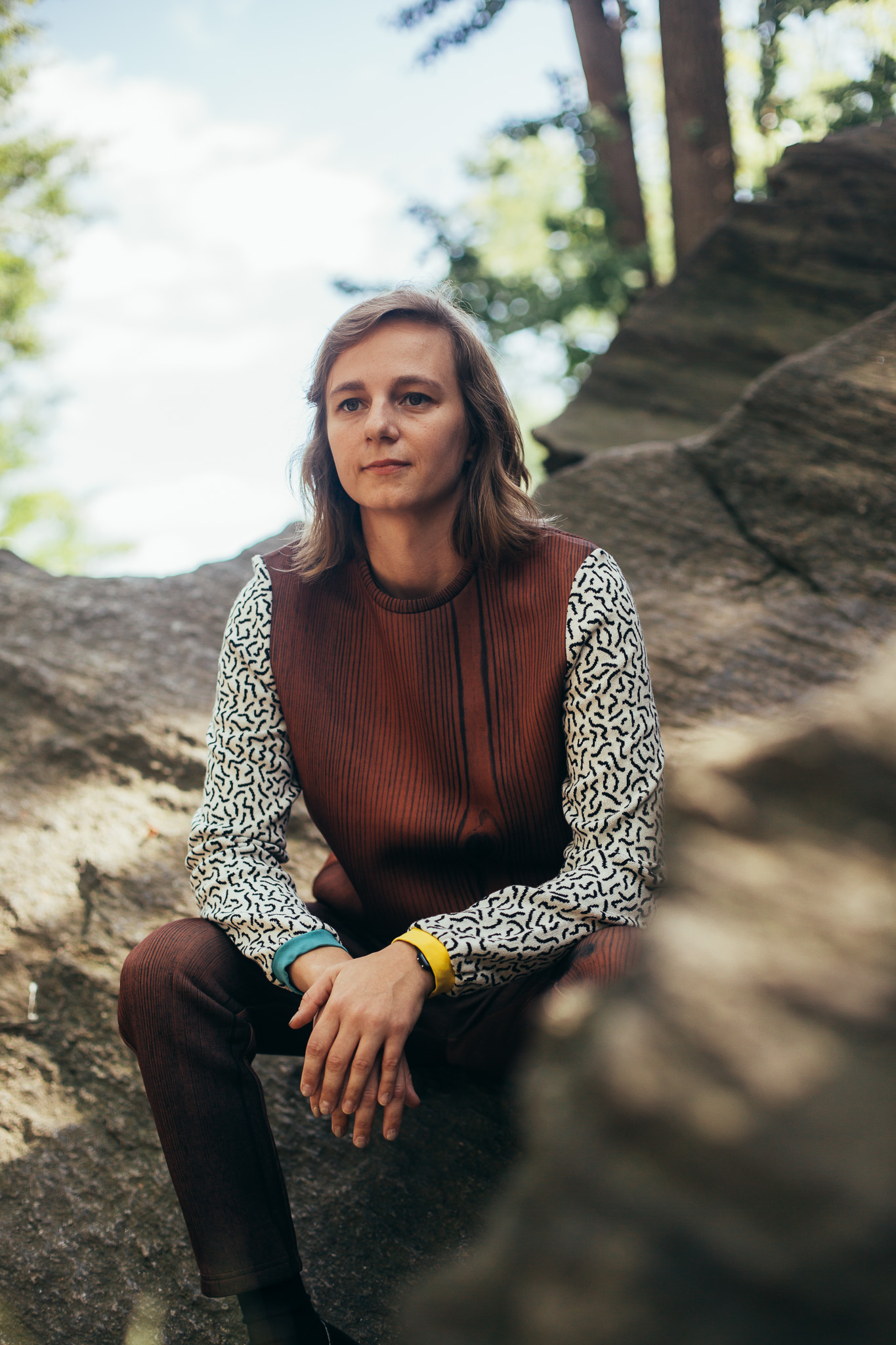 Portrait of Shannon sitting outside on a sunny day among large, flat rocks, smiling softly and looking off in another direction. She is wearing a wood grain sweatshirt and pant set. The sweatshirt has contrasting sleeves with a squiggle pattern and sleeve cuffs in two different colors.