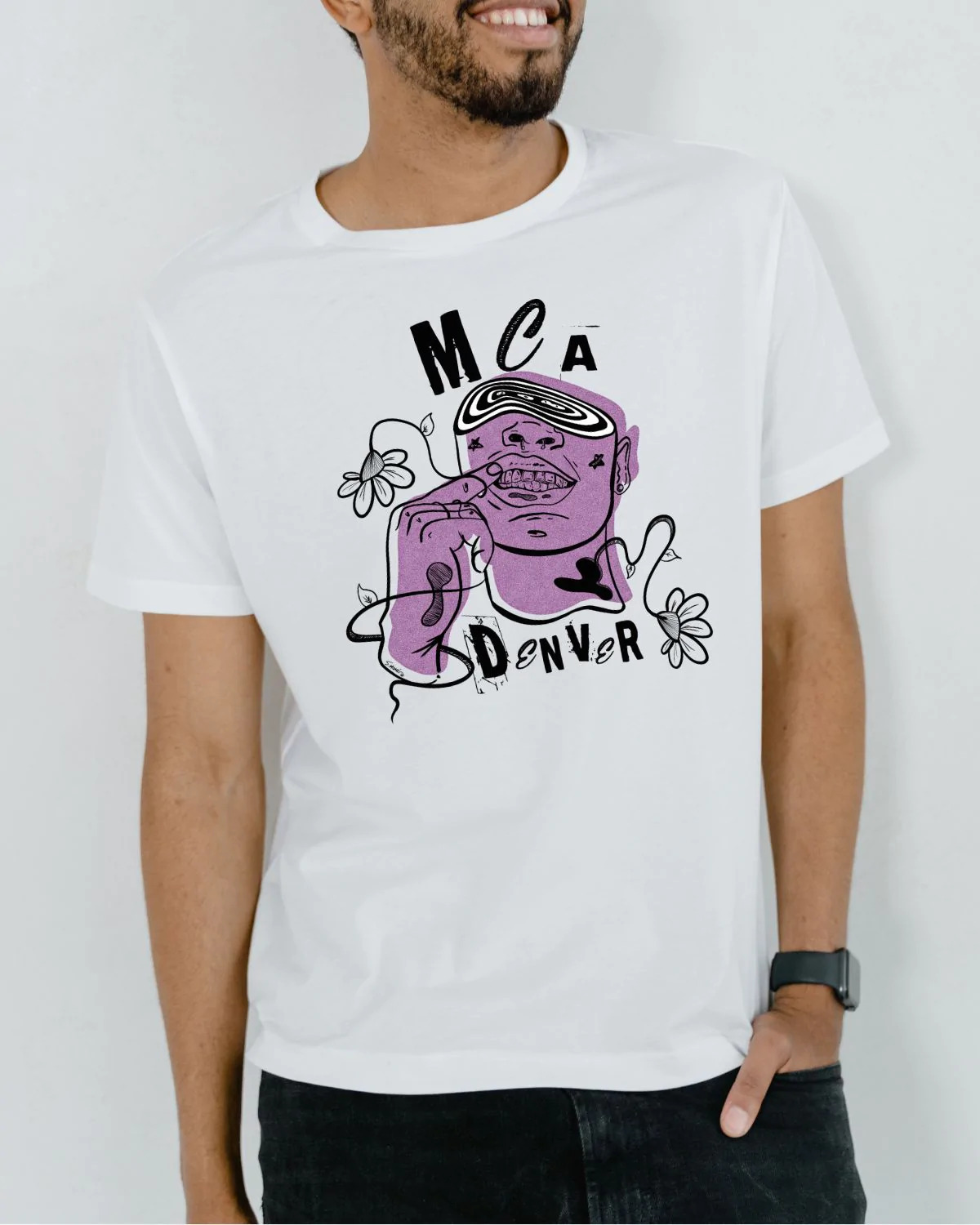 Model posing in front of a white wall and sporting a white t-shirt with a purple design on it. The t-shirt reads, “MCA Denver” and there is a head of a figure on it. The figure is sporting grills, there are flowers surrounding the figure, and there is a swirly vortex replacing where the figure’s eyes might have been.