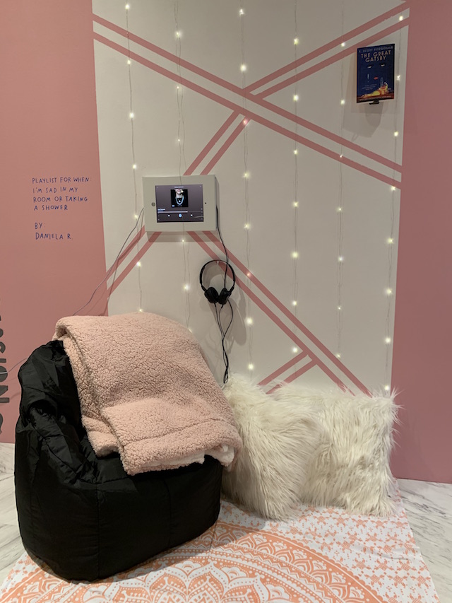 Close up of white triangles are painted onto the pink wall. String lights surround the pattern. A monitor and headphones are mounted on the wall. Text reads: acquelin Olivas - This playlist is for when I’m sad and when I’m with friends being in la depre