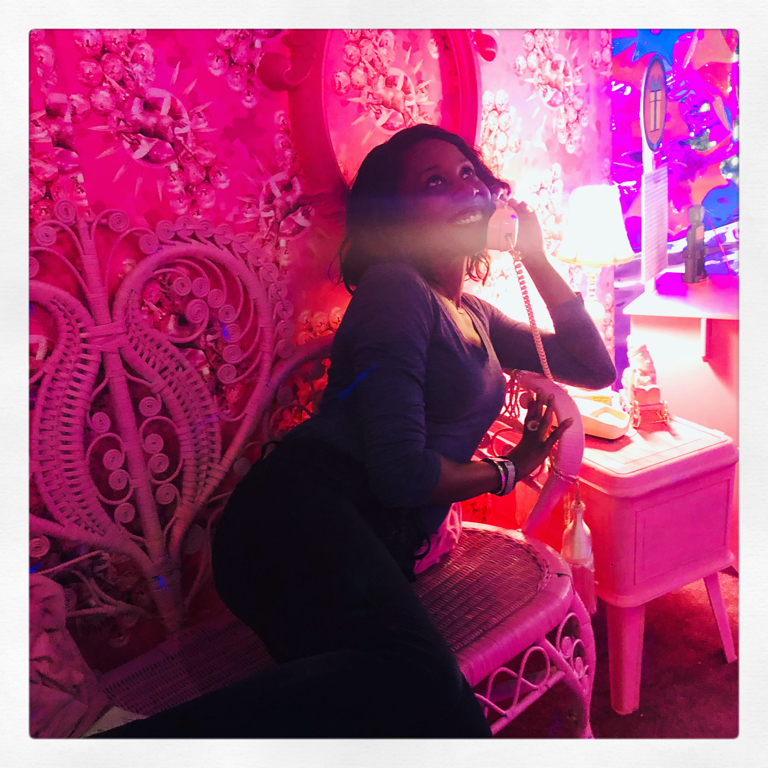 portrait of Tai in an eclectic, pink colored room. She’s lounging on a bench and is holding up a corded landline phone, looking up and smiling. 