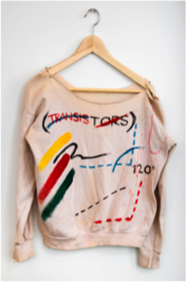 An off white colored sweater hangs on a wood clothes hanger on a white wall. It is hand painted with abstract shapes and the words TRANSISTORS is in red near the collar. 