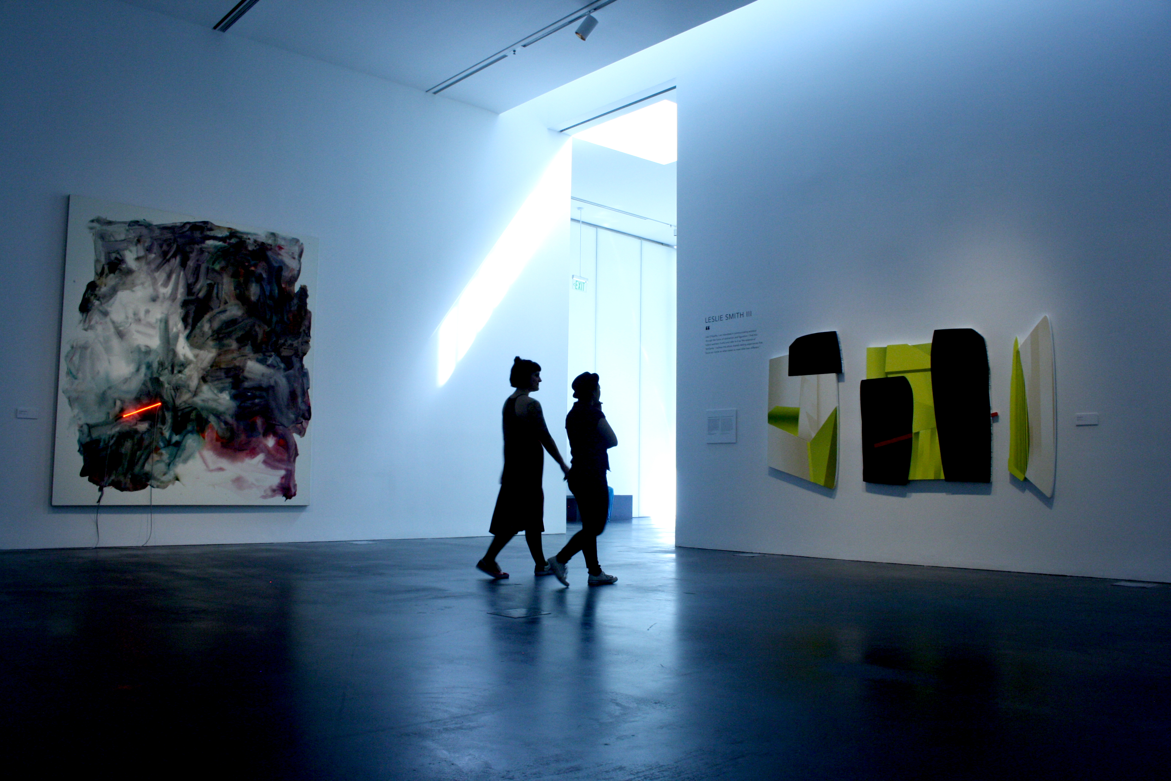 Two unidentifiable people in a gallery look at an abstract artwork. The ceiling is high and the floor is concrete. 