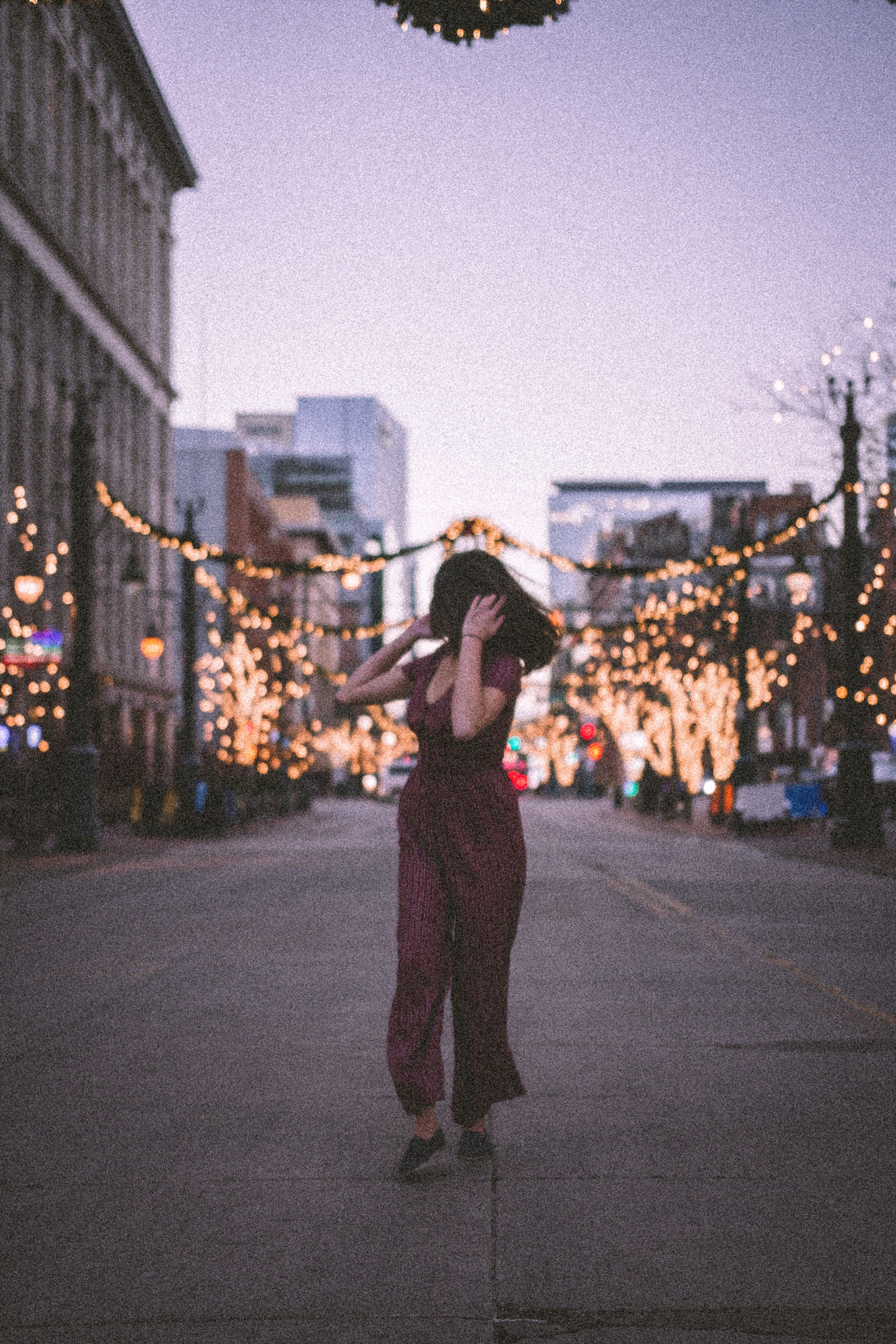 A young woman walks down the middle of a street at dusk. She looks behind her shoulder at holiday lights that adorn the street lamps and trees. 