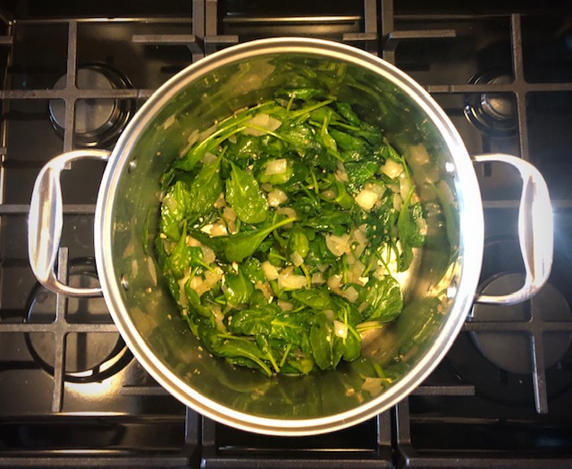 Birds eye view of spinach and other ingredients in a stainless steel pot on a gas range. 