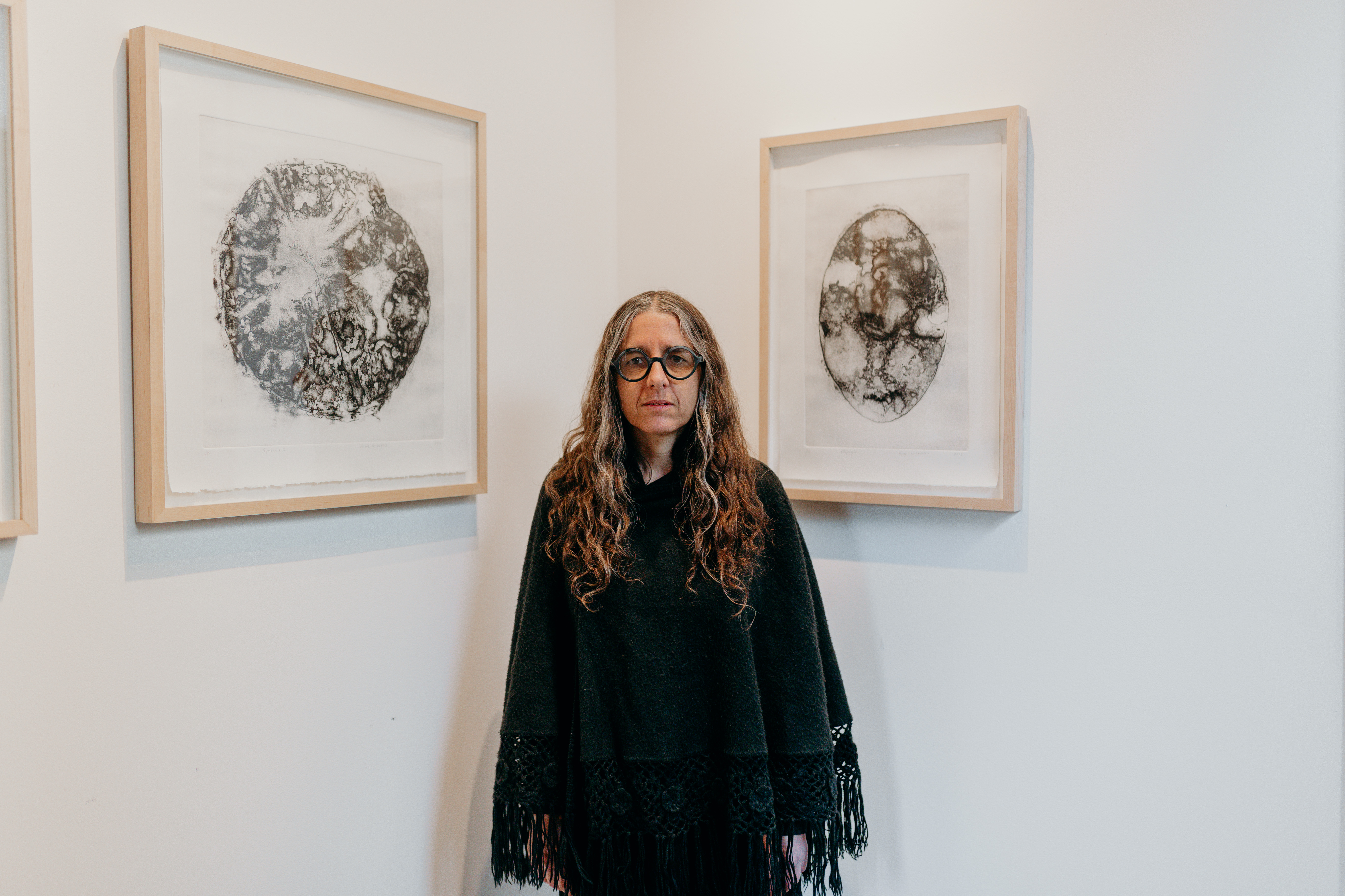 An older woman in a black shawl poses in front of two black and white artworks. They resemble stones, or even a brain. 
