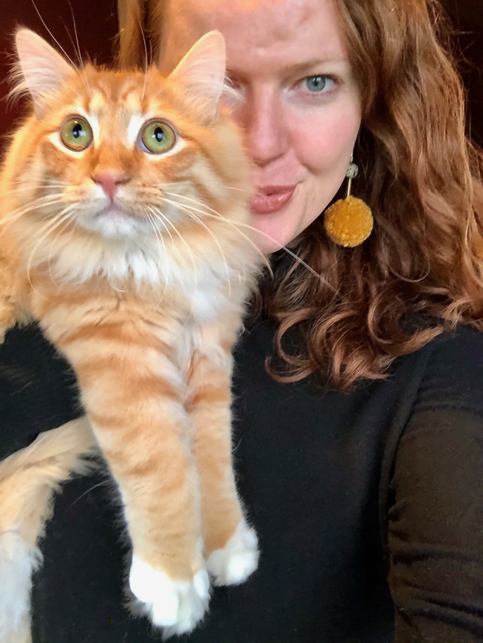  selfie of Krista with her cat. She is piercing her lips and is wearing a black turtle neck, yellow pom-pom earrings, and her red short hair is in curls. Her cat’s coloring is orange and white, and the cat has green eyes, which appear aghast. 