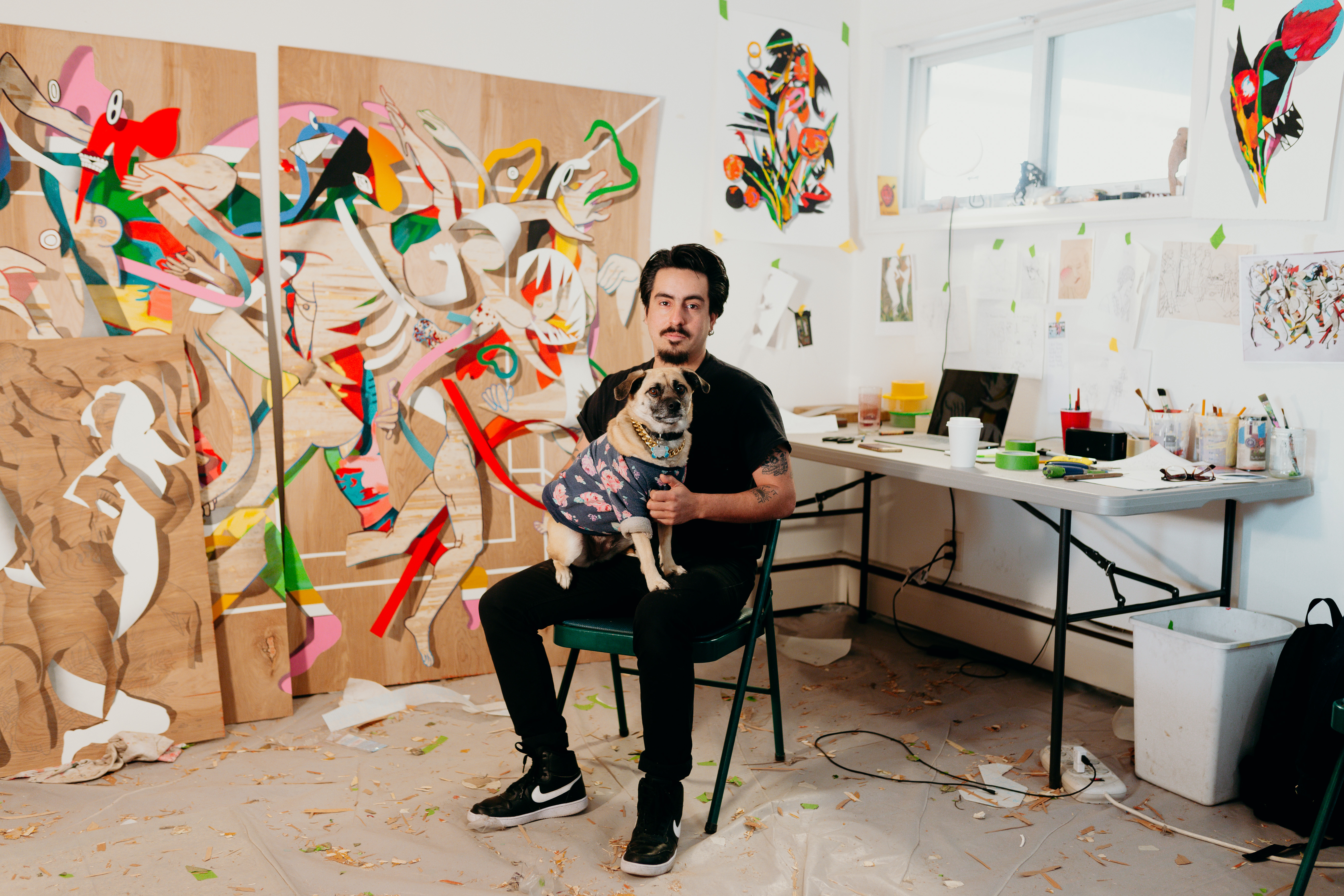 A man sits in a chair with a small dog in his lap in a studio space with  large paintings resting against the walls. 