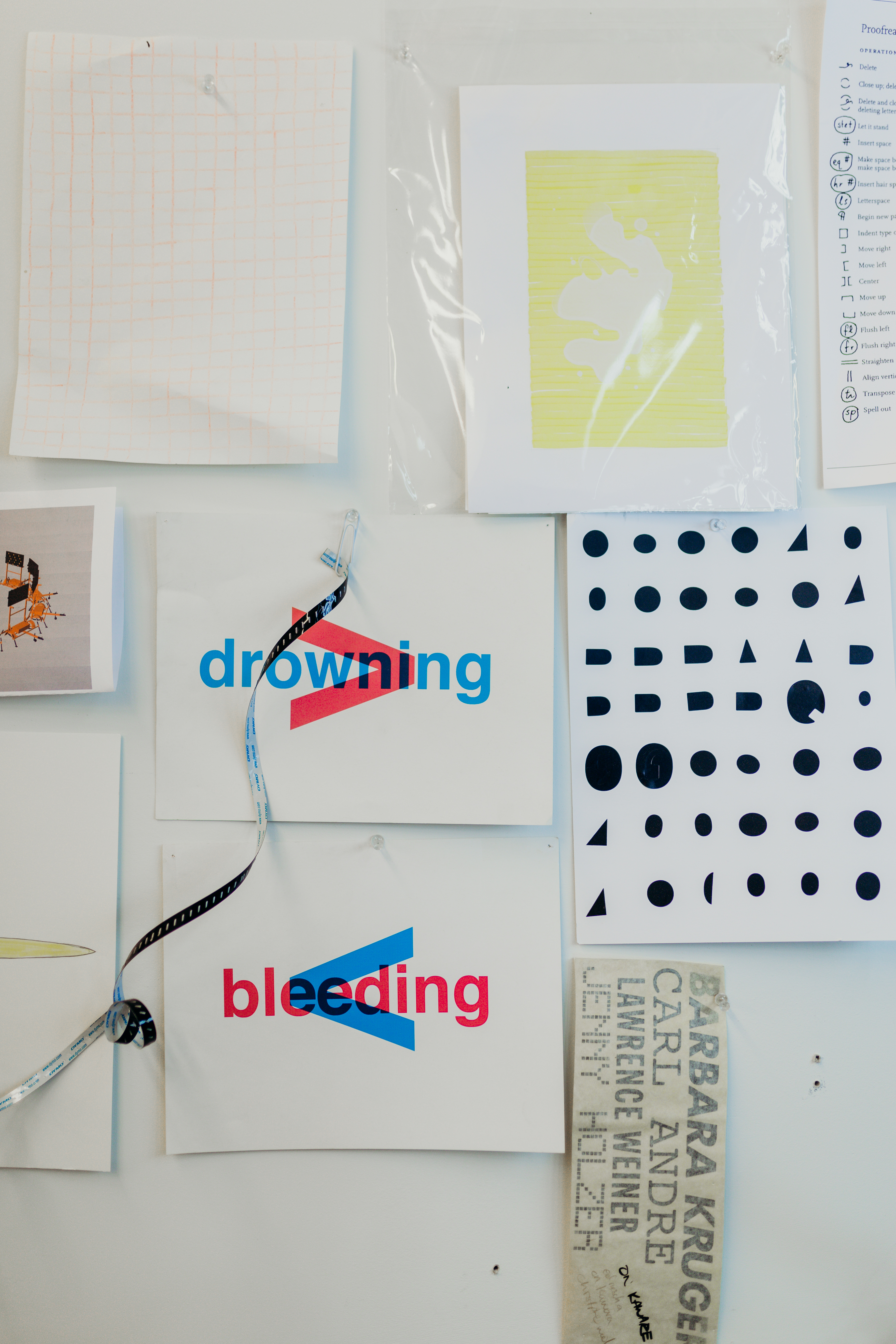 Artworks hang on a white wall. The words bleeding and drowning are superimposed over the grater than and lesser than symbols in blue and red. 