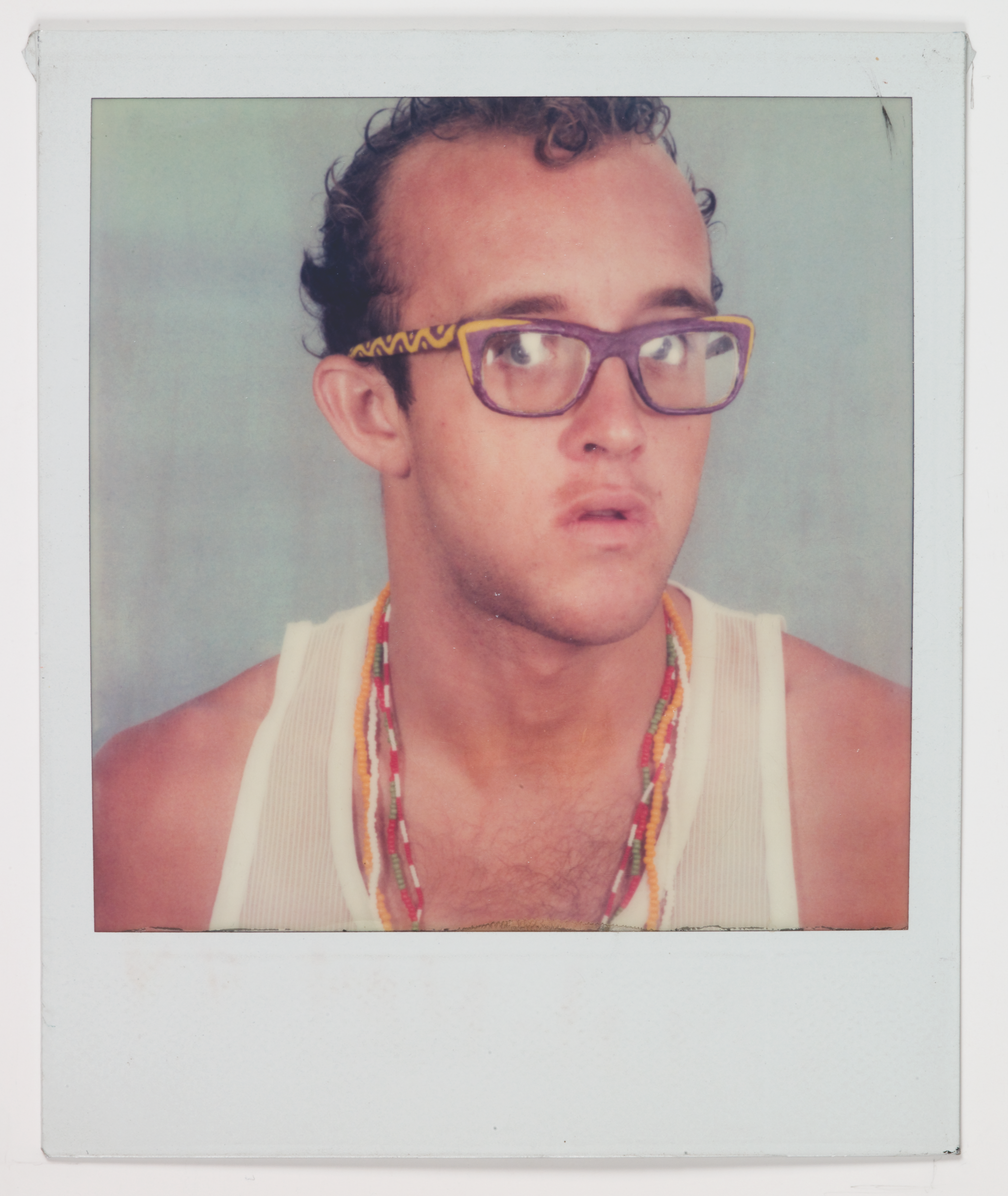 Image of artist Keith Haring. He wears purple glasses with yellow designs on the sides, a white tank top, colorful necklaces, and has short curly hair.
