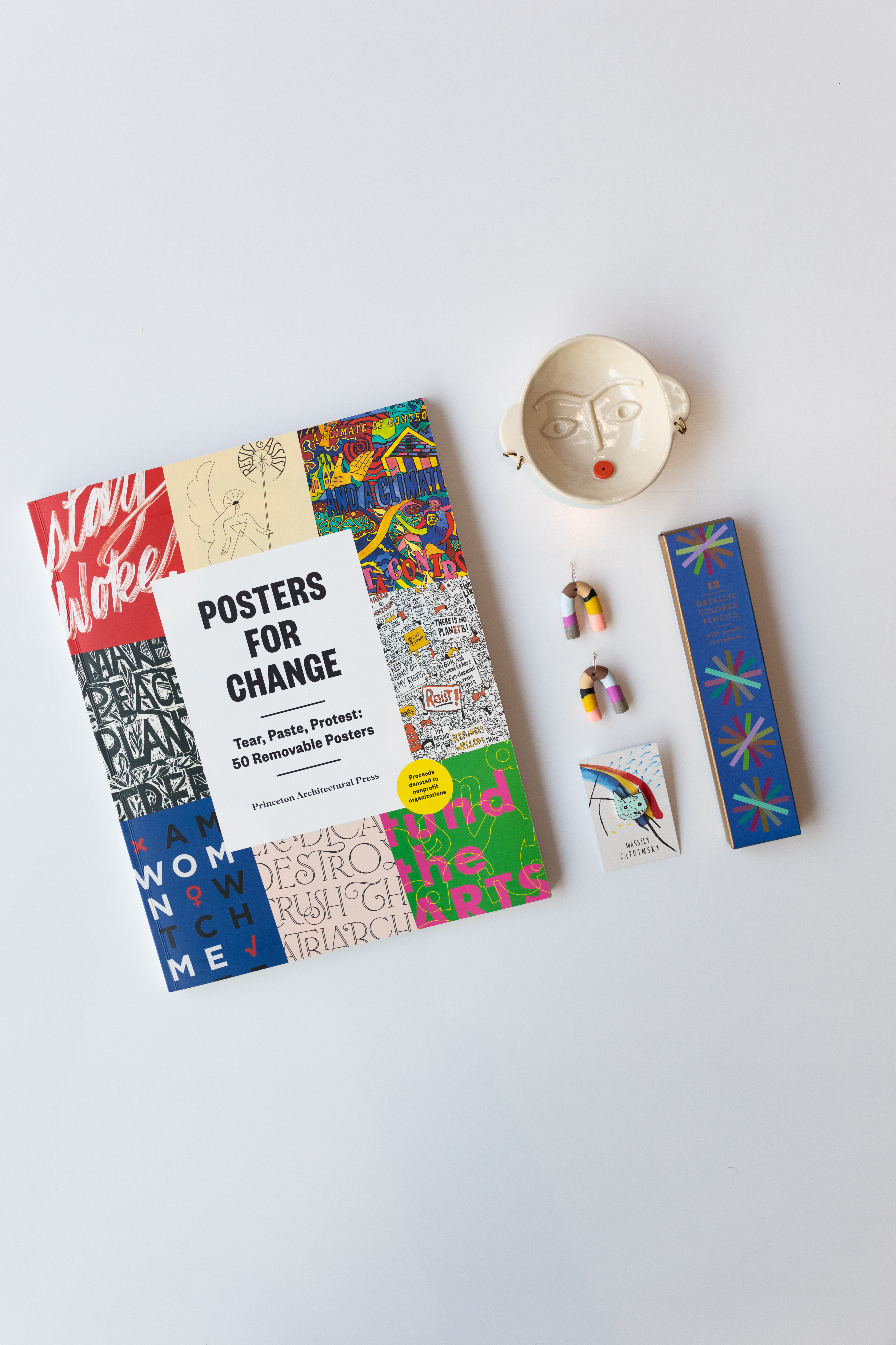 A book titled posters for change; colorful earrings; a jewlery holder with facial features on the bottom;  a cat in the style of Wassily Kandinsky, appropriately titled Wassily Catdinksy; a blue box containing 12 metallic colored pencils. 