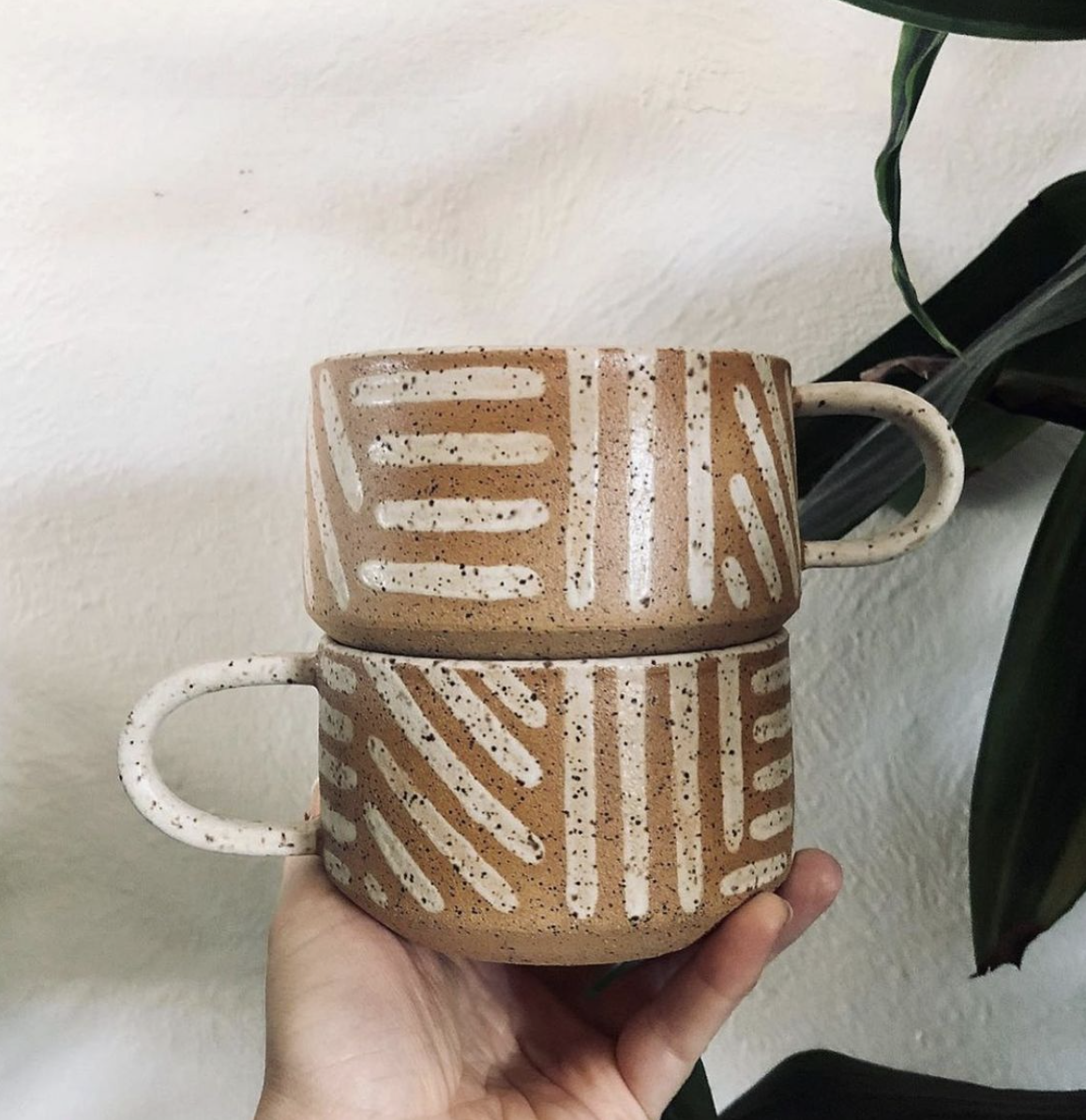 Two small mugs with round handles stacked on top of each other and held in one hand. The mugs are light brown with black speckles on them, adorned with lines of cream brush strokes going in different directions. The mugs are held up against a white textured wall and there is a large green plant on the left side of the frame. 