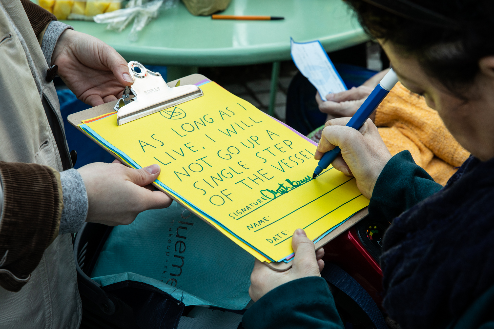 Close up photo of someone's hands holding a clipboard for another person to sign the pledge that states, "As long as I live, I will not go up a single step of the Vessel" in green text written on a piece of bright yellow paper.