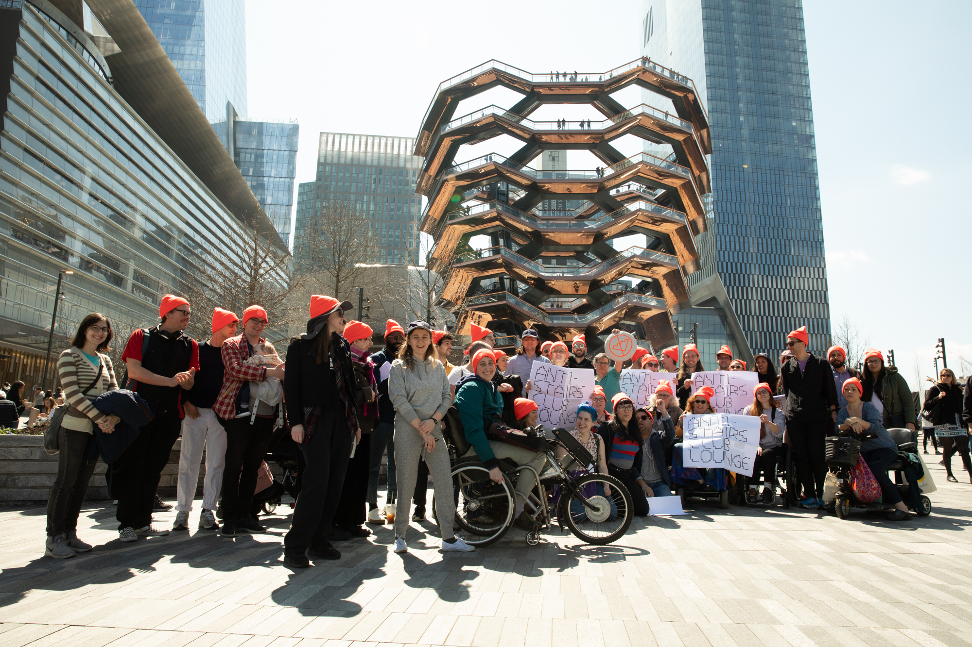 A group photo of people at Hudson Yards in Manhattan in front of the ‘Vessel’ — a building-sized, basket-like structure made of 154 interconnected stairways. People in the group are wearing bright orange Anti-Stairs Club Lounge beanies and holding Anti-Stairs Club Lounge signs.