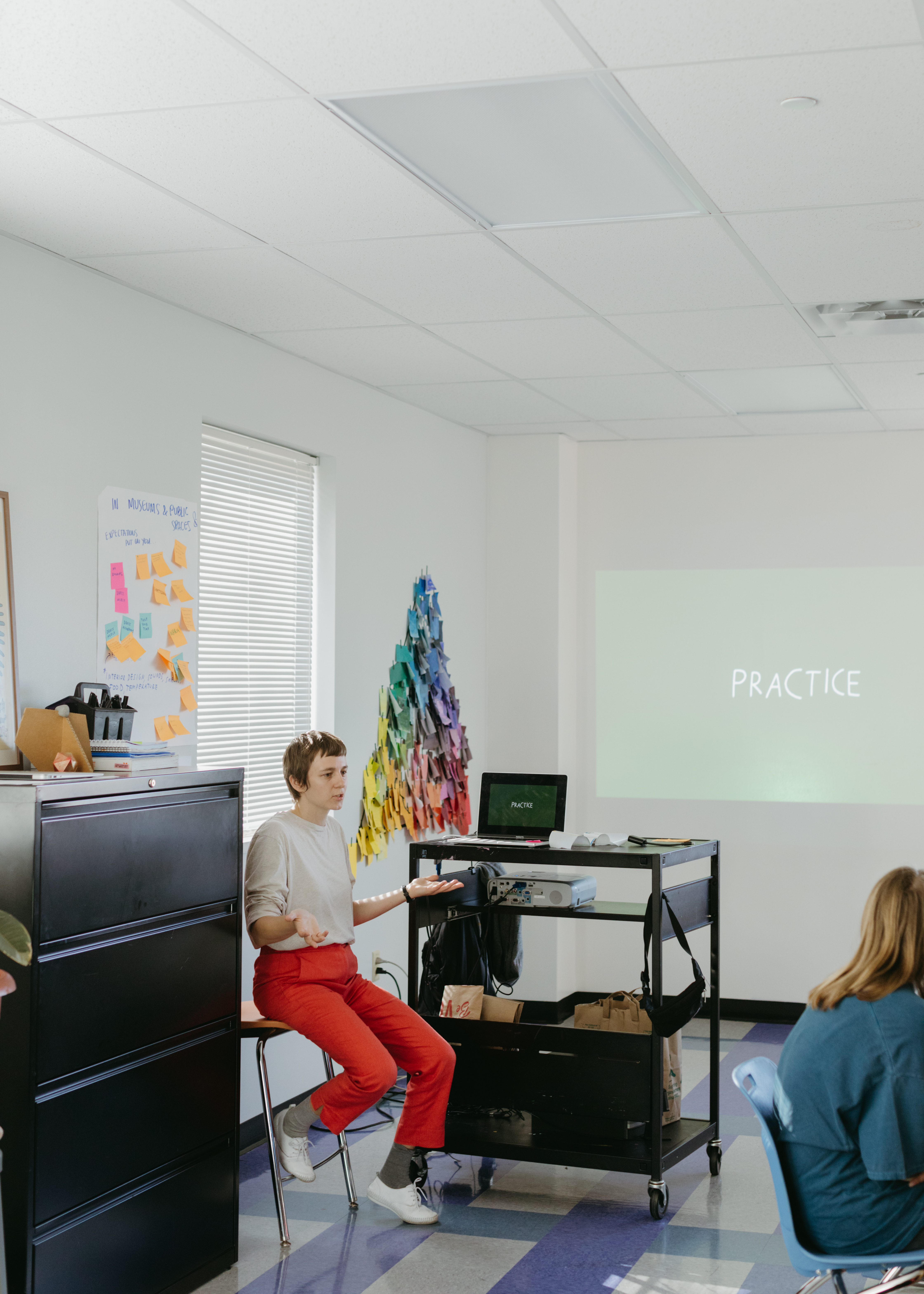 A photo of Shannon at Girls Athletic and Leadership School (GALS), giving a presentation, with the word "Practice" being projected onto the wall. She is sitting in a chair next to the presentation, talking with her palms facing upwards, wearing a beige top and bright orangish, pinkish pants.