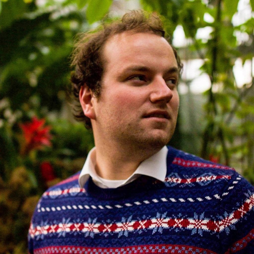  portrait of Brad in front of what looks like a lush green plants. His hair looks unkept but cool, and he is looking off into the distance with a contemplative, serious look on his face, and is wearing a festive blue and red sweater. 