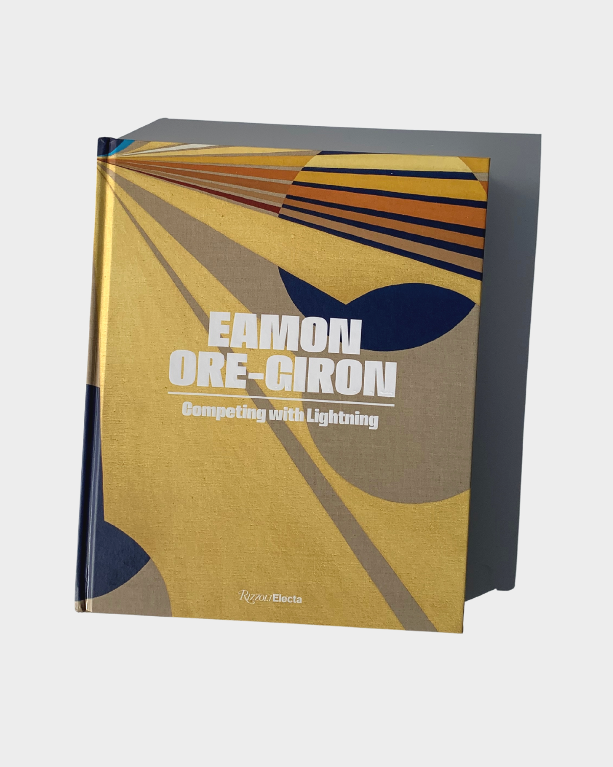 Exhibition catalogue photographed on a white background under harsh sunlight that creates a shadow. The white, bold text on the book reads, “Eamon Ore-Giron” and “Competing with Lightning.” The catalogue cover art is made up of geometric shapes in a primarily glittering gold color. 