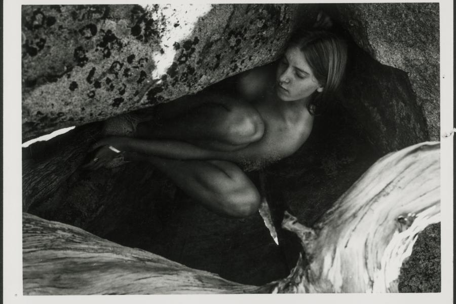 A nude woman is lying in the fetal position surrounded by rocks in black and white. She lies her head upon a stone with her eyes closed. She appears to be sleeping peacefully. 