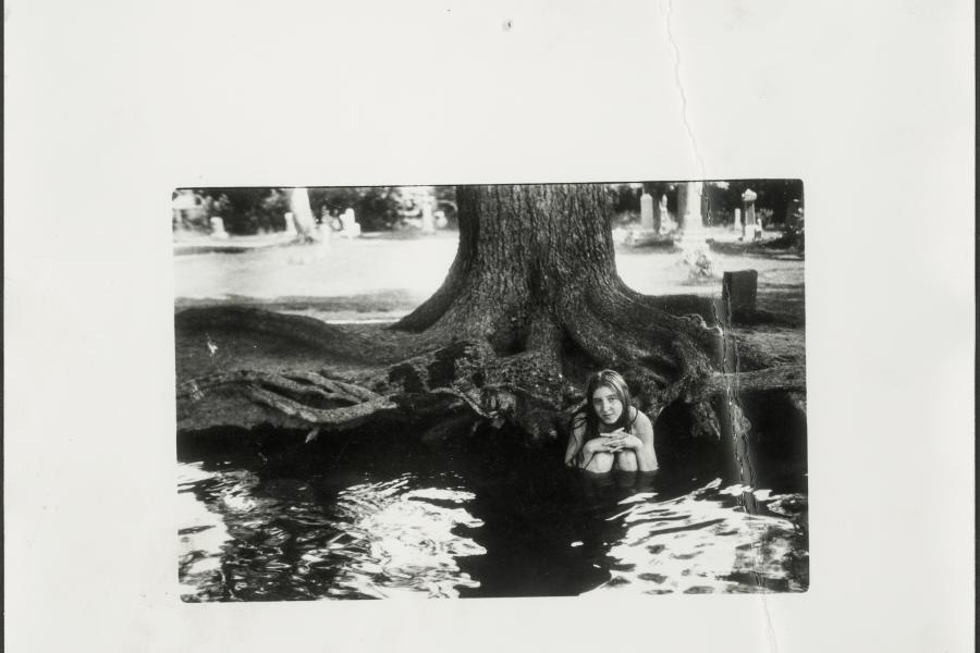  Francesca Woodman, Untitled photograph, circa 1975-1978. Gelatin silver print.  A woman sits in a pond with the base of a tree visible behind her. She has her knees to her chest and rests her hands upon her knees. She looks into the camera with a longing look on her face. 