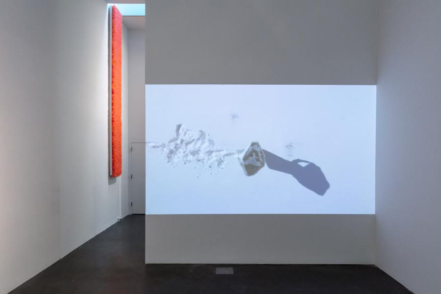 MCA Denver gallery showing a video projection on a wall. The video shows a bird's eye view of a person carrying a boulder in the snow.