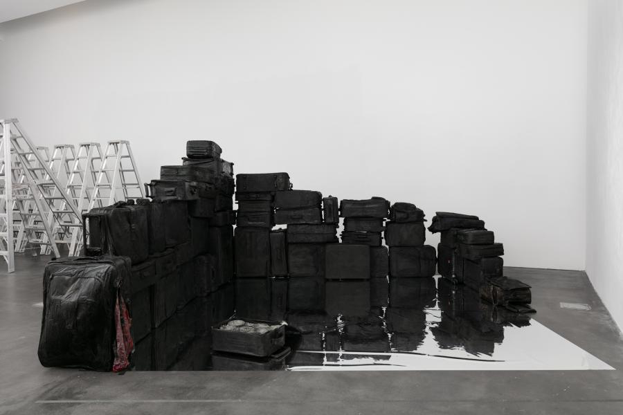 Black suitcases stacked on top of one another in an MCA Denver gallery. Next to the suitcases is a large puddle of water. 