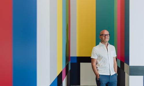 A portrait of former director Adam Lerner poses in front of a wall painted to resemble technicolor on a tv set. He is bald, wearing a white short sleeve button up, and blue chinos. He smiles gently. 