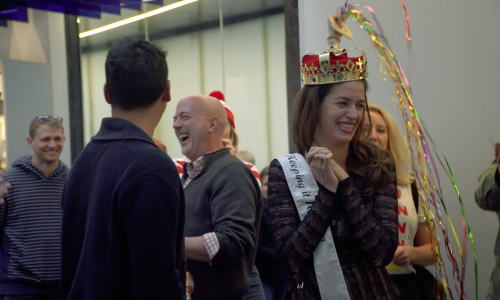 A man and a woman are surrounded by several people. The woman is wearing a white sash and a toy crown. 