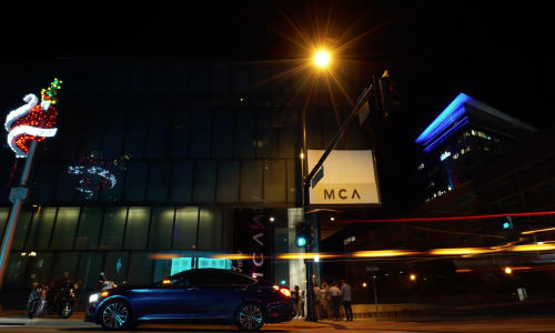 Exterior shot of the MCA Denver at night from street view. A heart sculpture is lit up and several people wait near the entrance in a line. 