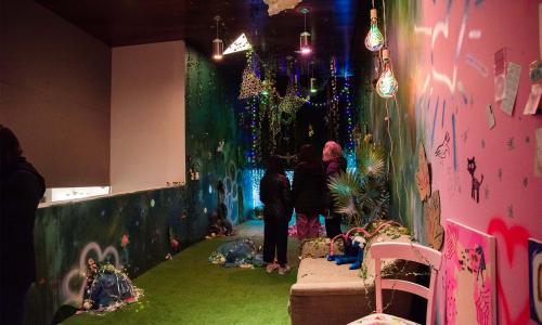 Gallery space featuring a magical immersive installation that is bright pink and deep forest green. There are little colorful pom poms and string lights affixed to the wall, along with other youthful illustrations and clippings. Three visitors stand in the center of the space near a tropical plant and a couch. There are vines and streamer-like material hanging from the ceiling. 