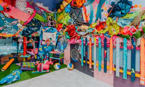  Teen installation featuring various materials and collage, including a colorful picket fence and a variety of colored fabrics attached to a purple and pink wall.