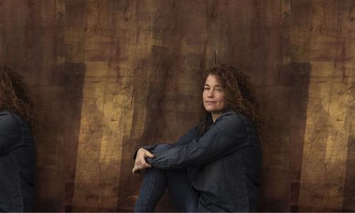 Portrait of street artist Swoon, posing in front of a brown textured backdrop and sporting a soft smile and curly hair.