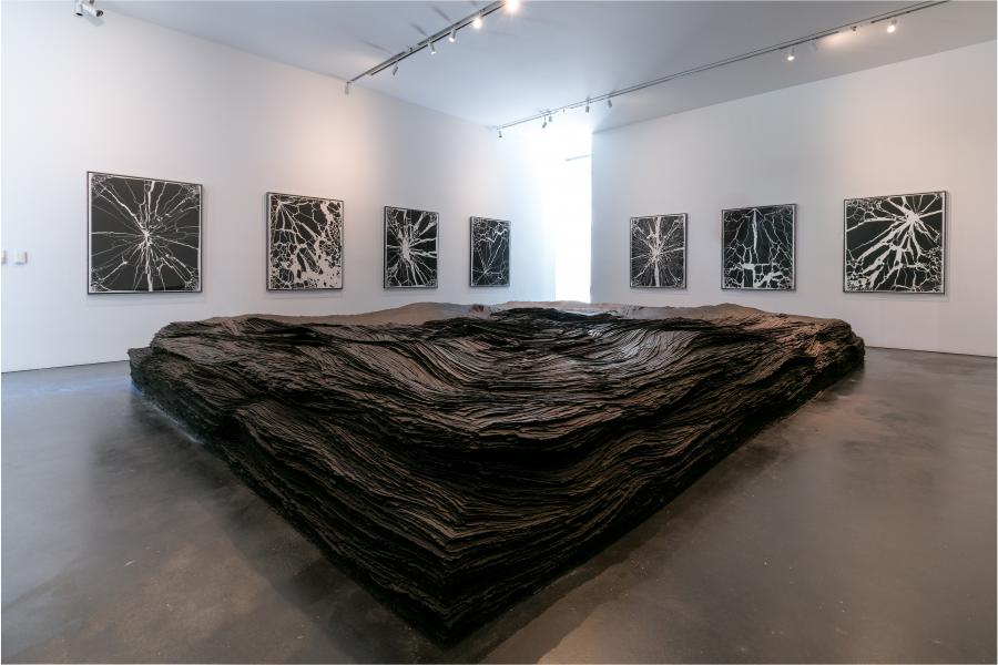 A large mound of black tar paper rests in the center of a gallery space. It resembles a charred earth. 