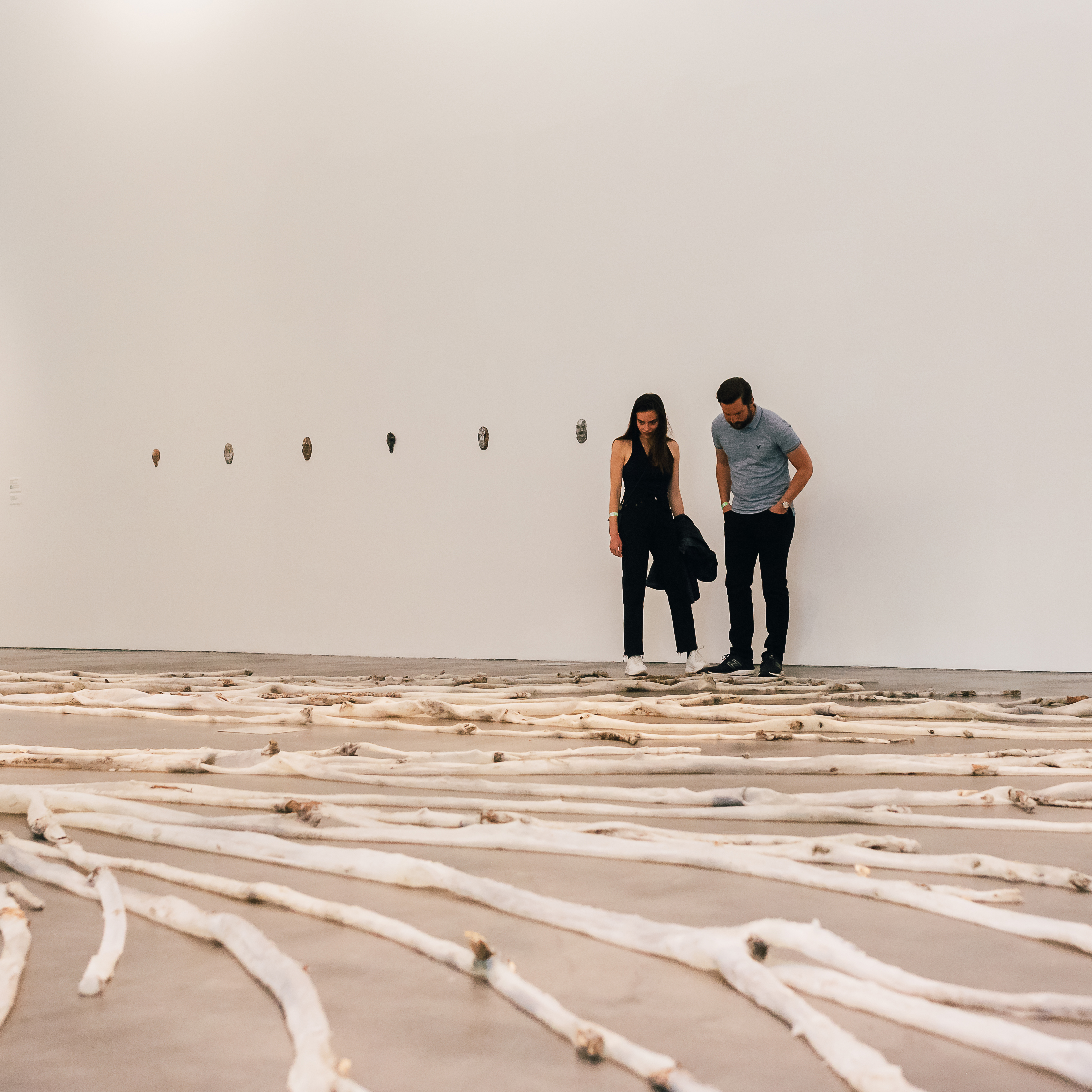 Two visitors looking down at a large plastered tree sprawled out on the ground in a gallery with white walls and dark flooring.