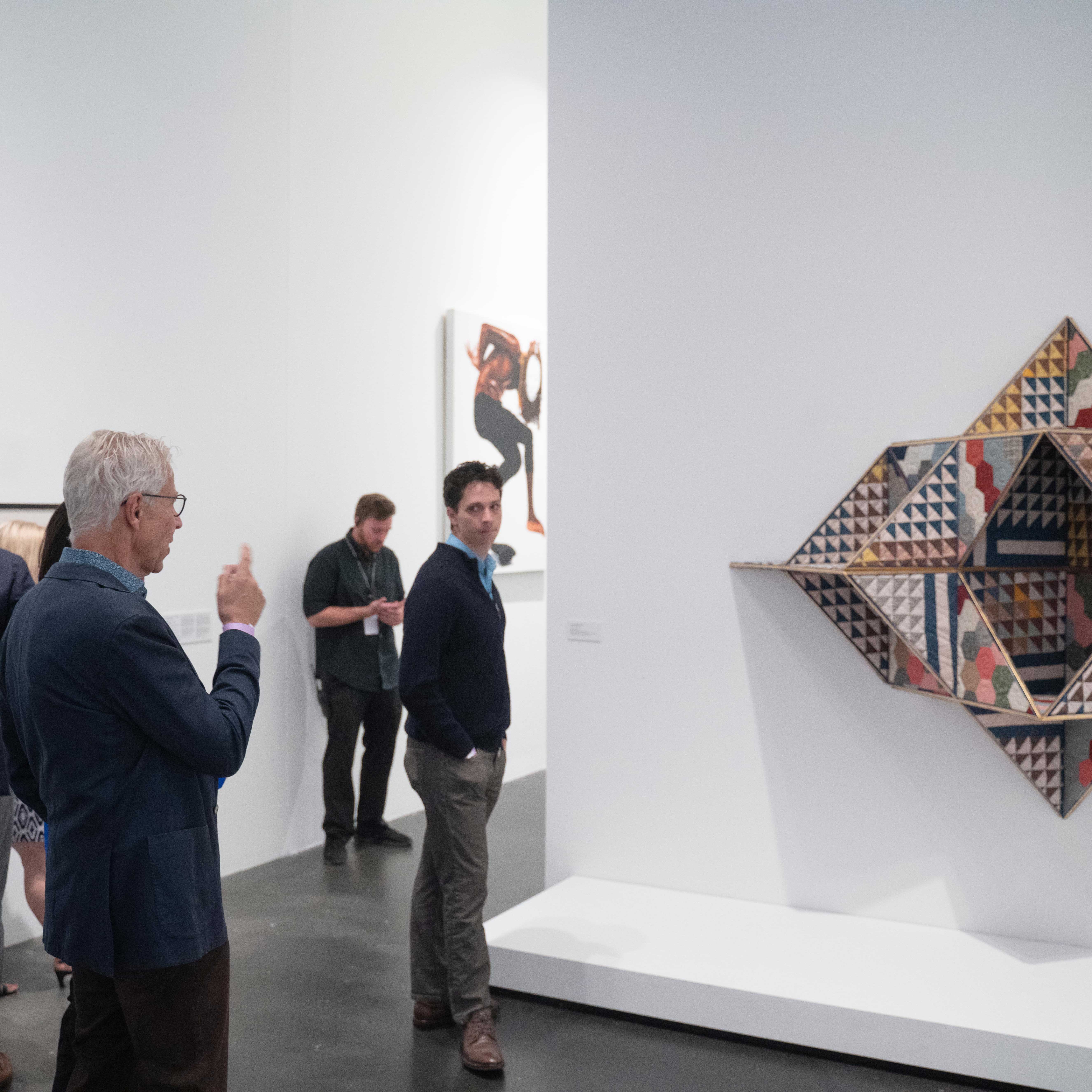 Visitors standing near a quilt that it's a geometric shape and affixed to the wall. 