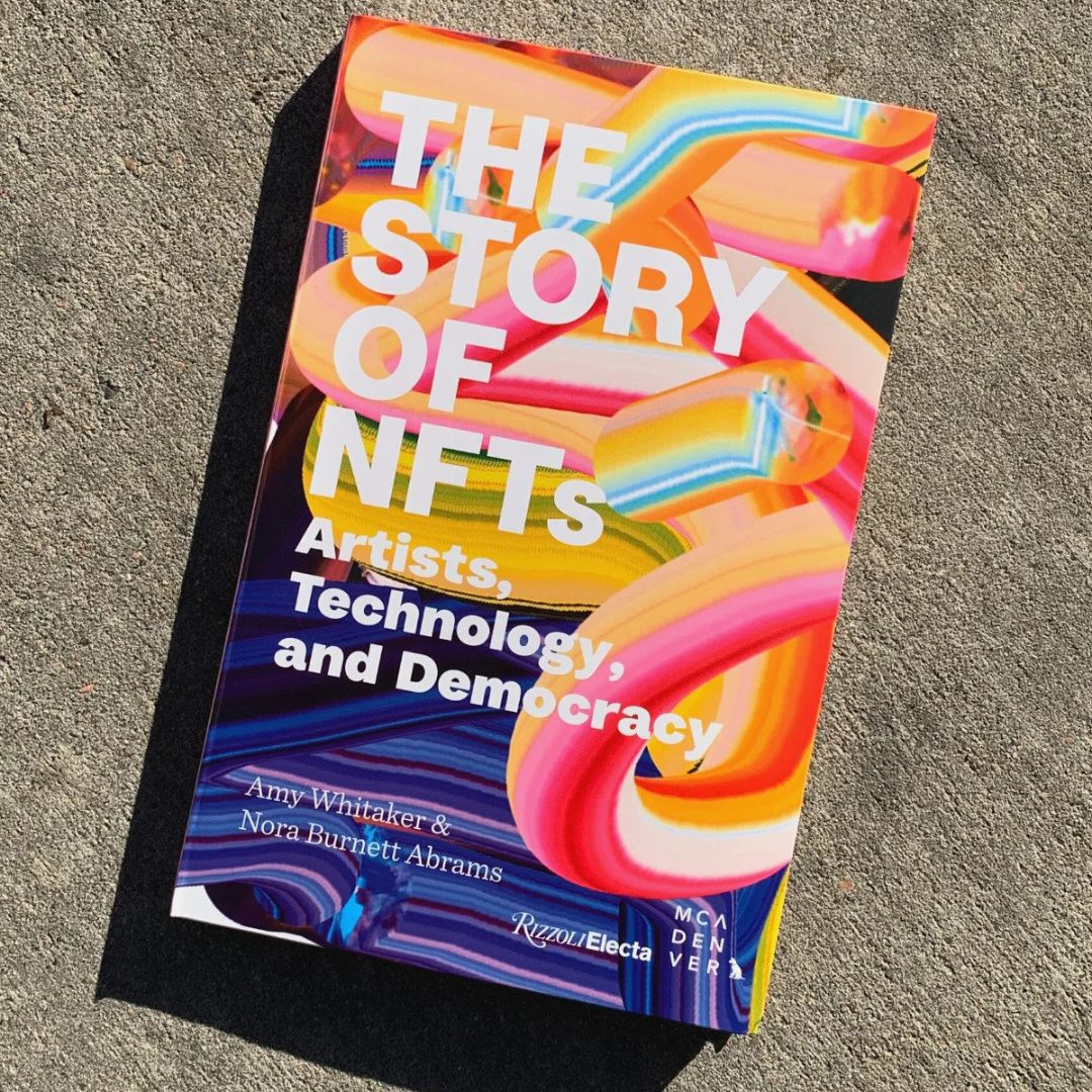  Book photographed on concrete in bright sunlight. The title of the book, printed in white font, reads “The Story of NFTs”. The cover of the book is colorful and looks like layered brush strokes.