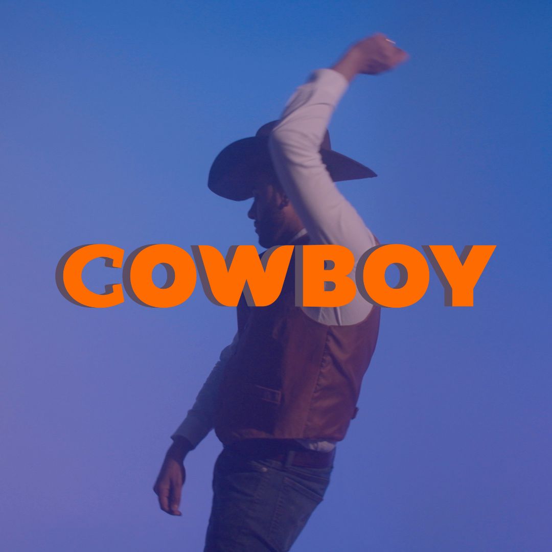 Still from a video of a cowboy against a bluish, purplish sky. The cowboy is sporting a cowboy hat and vest, and has one arm up in the air.