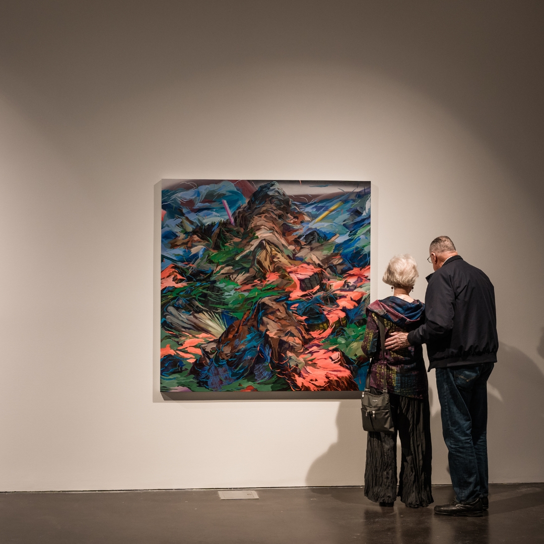 Two people with their backs facing the camera, looking at a colorful painting on the wall.