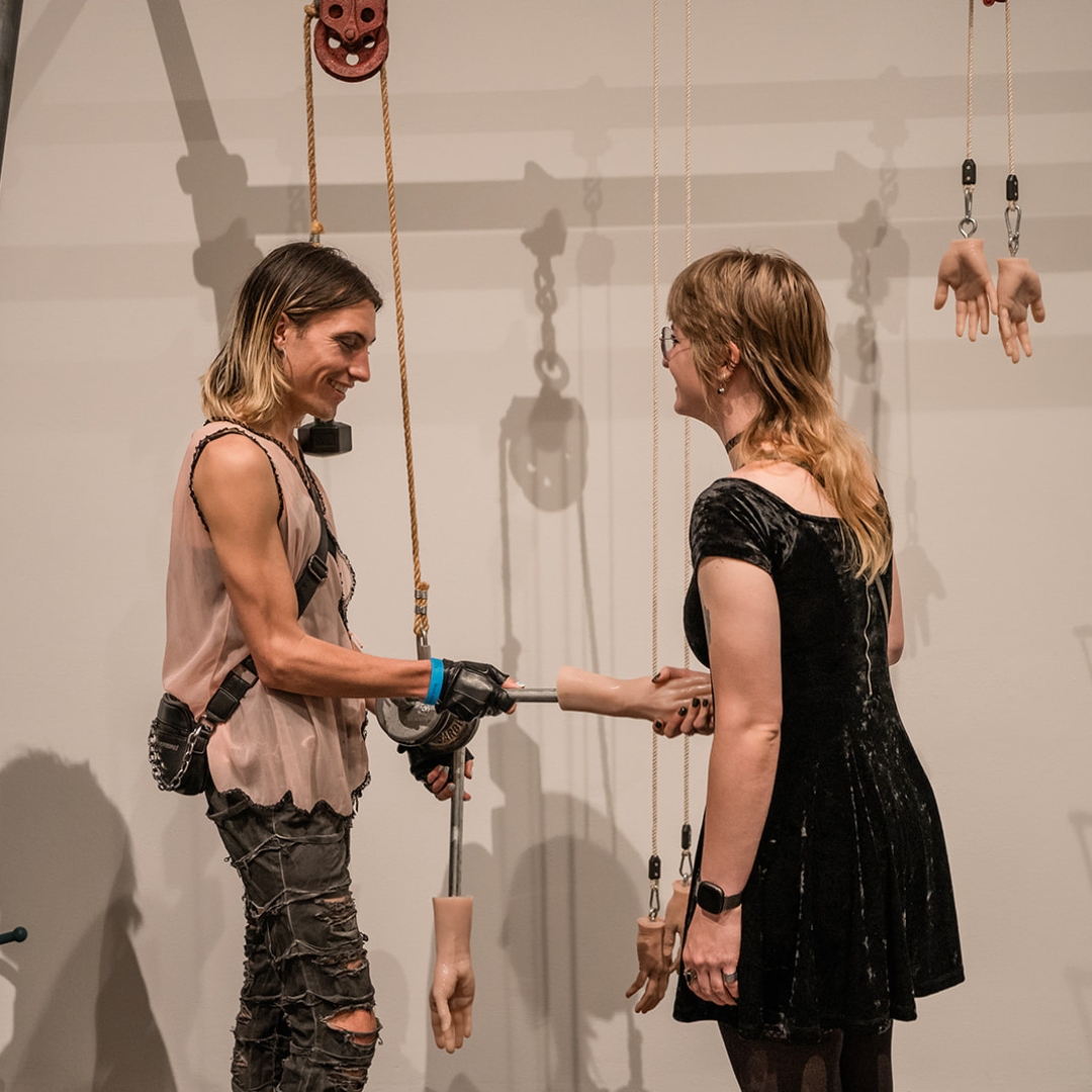 Two people in a gallery holding a hand that is a part of an art installation.