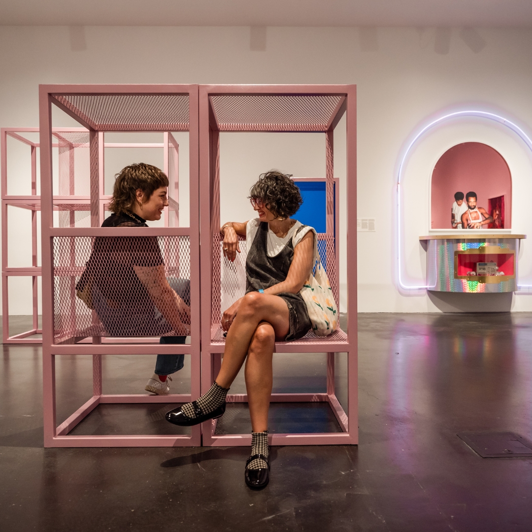 Two people sitting in a gallery on a light pink sculpture.