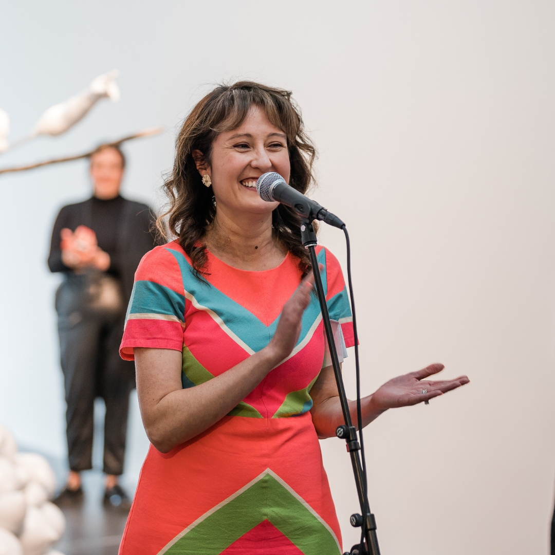 Curator Miranda Lash smiling in front of a microphone in a gallery.