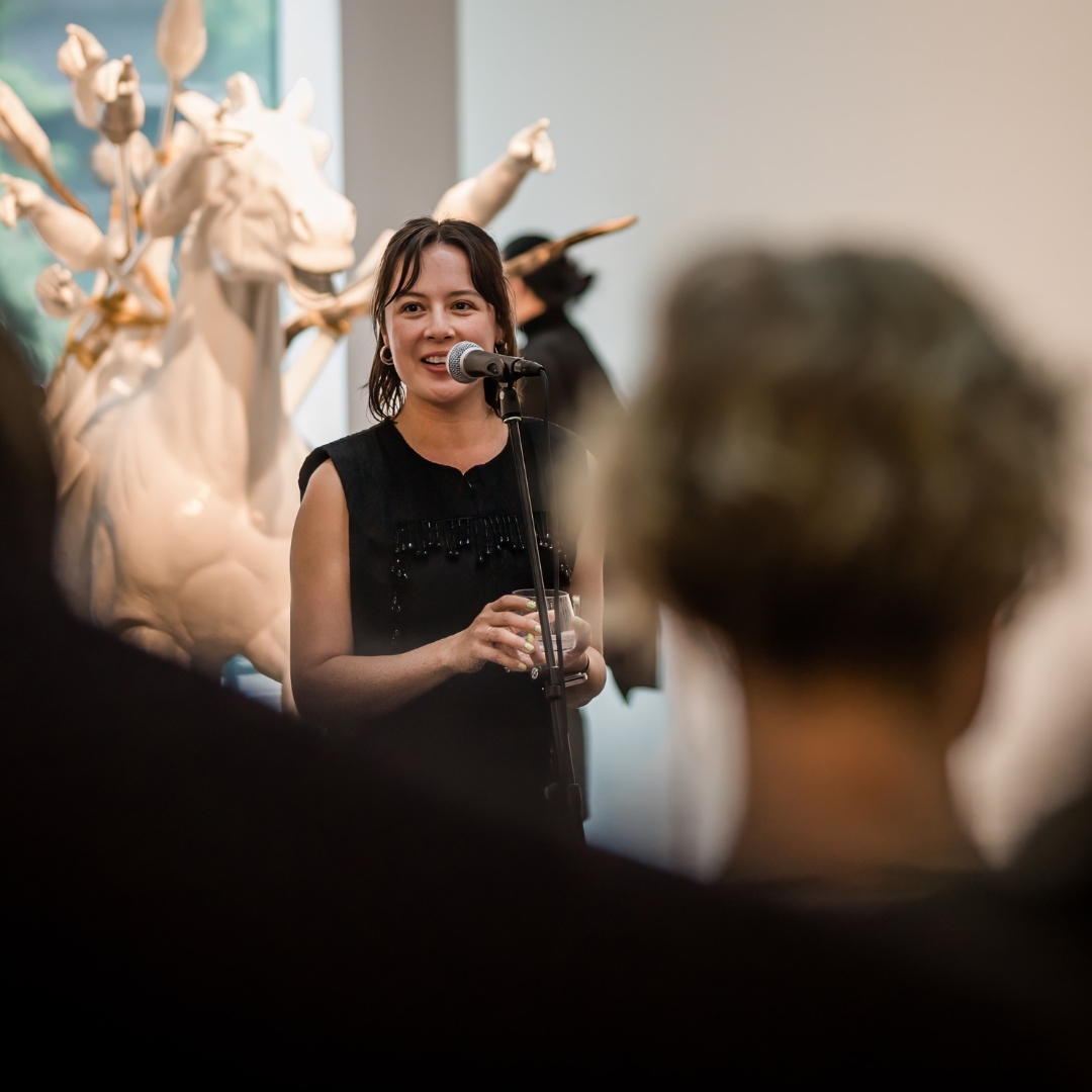 Curator Leilani Lynch smiling in front of a microphone in a gallery.