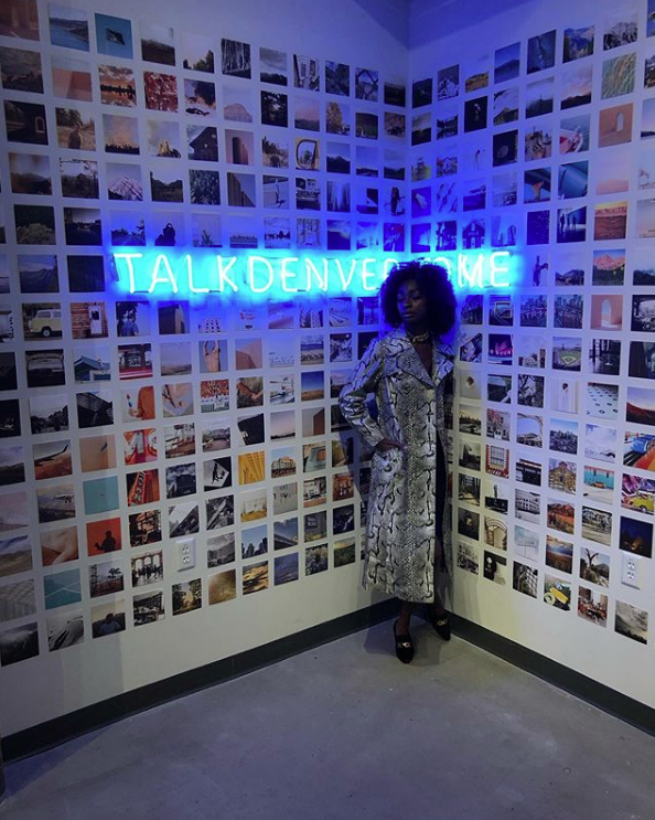 A woman in a snakeskin trenchcoat poses in front of a wall covered in photographs. There is a neon sign in blue that reads Talk Denver to Me. 