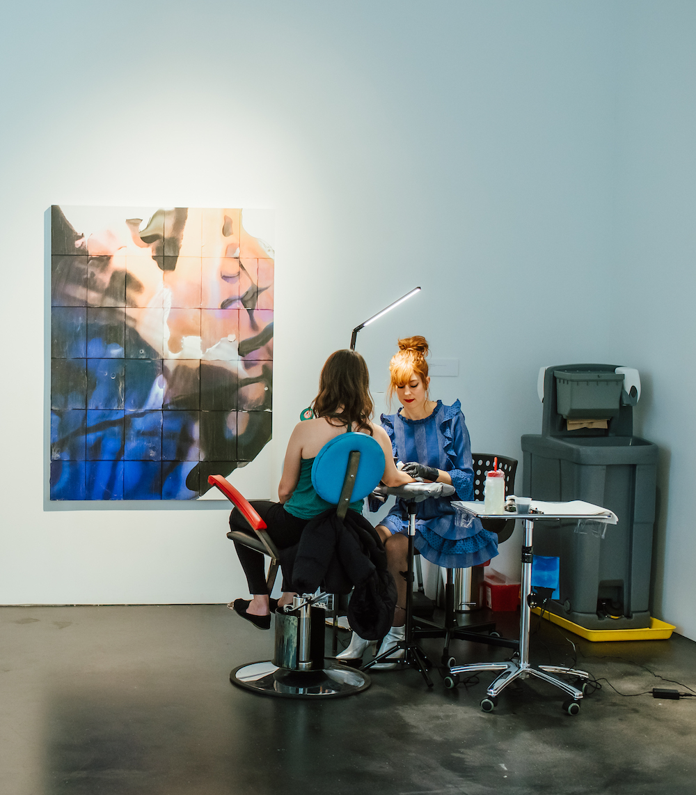 A woman tattoos another woman in a gallery space. There is an abstract artwork on the wall behind them in pink, beige, black, and blue shades that seems aquatic.