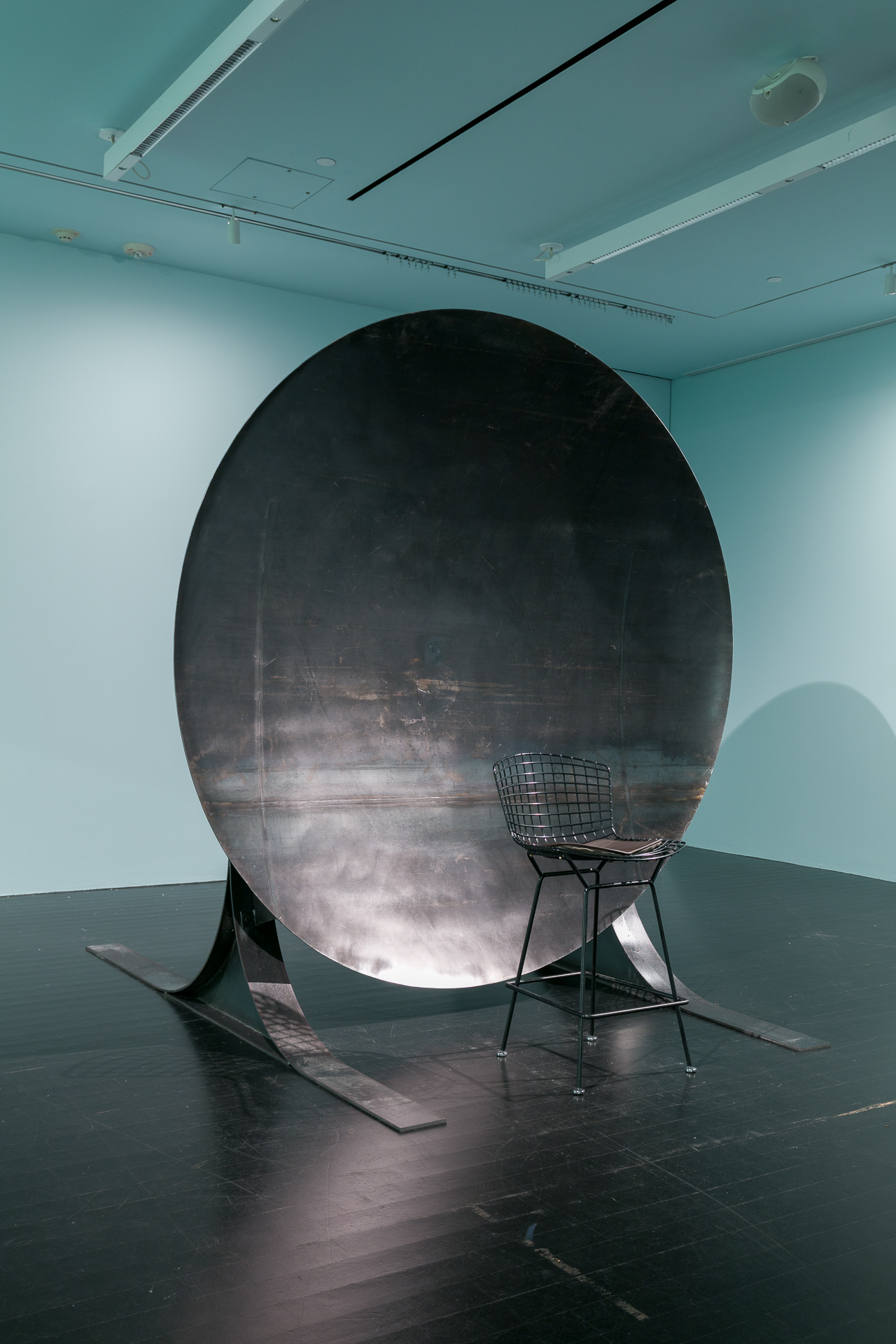 [Image description: An image of an art installation in a white gallery space. The work consists of two chairs, each sitting in front of their own large discs, which are meant to replicate the effects of an acoustic mirror. The discs reflect and focus sound waves in a way that visitors can sit far apart but will hear each other as if they are standing right next to each other.]