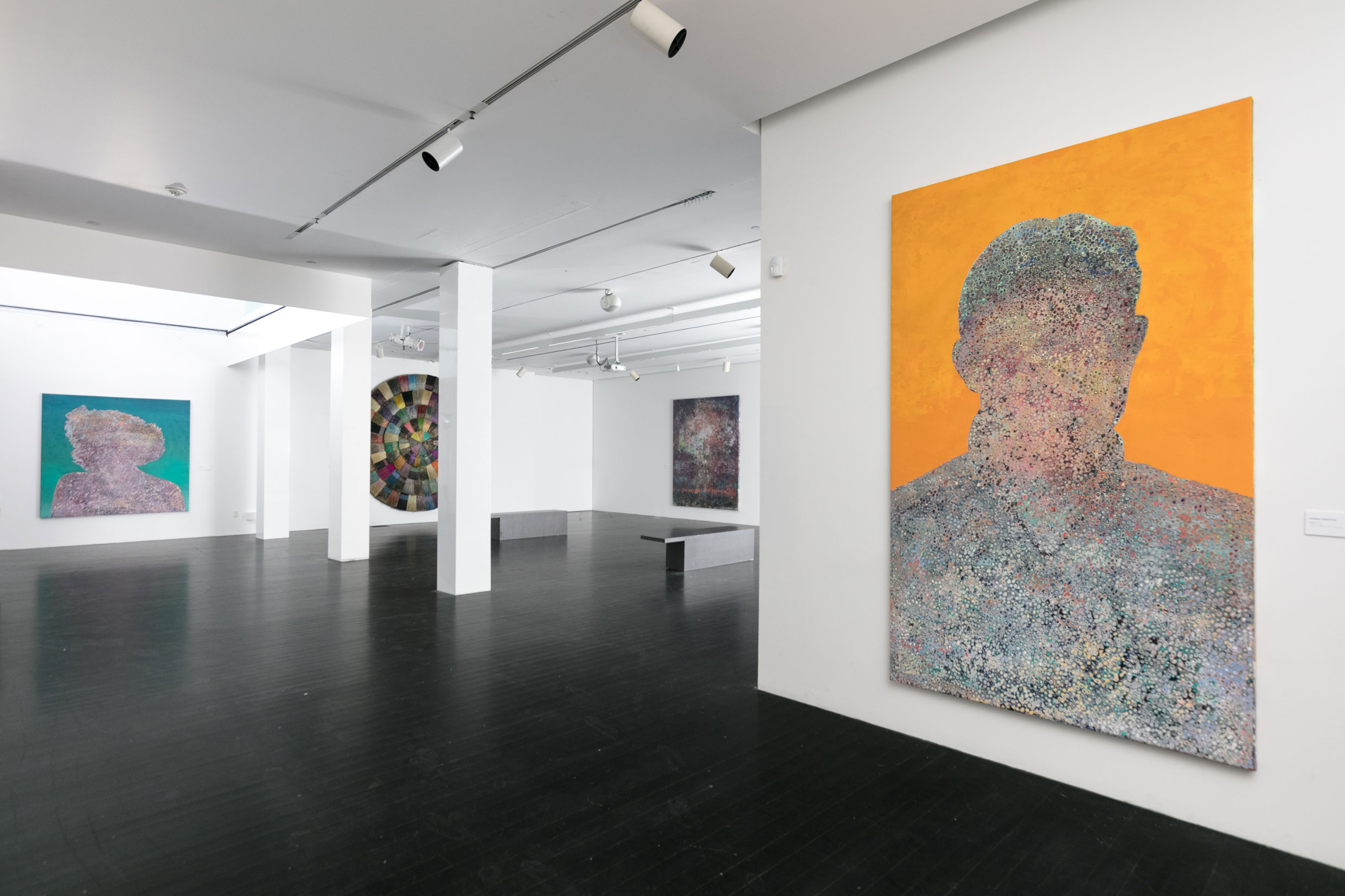 A gallery space filled with large paintings hanging on white walls. The artworks are abstract depictions of unidentifiable people. Almost like a silhouette. 