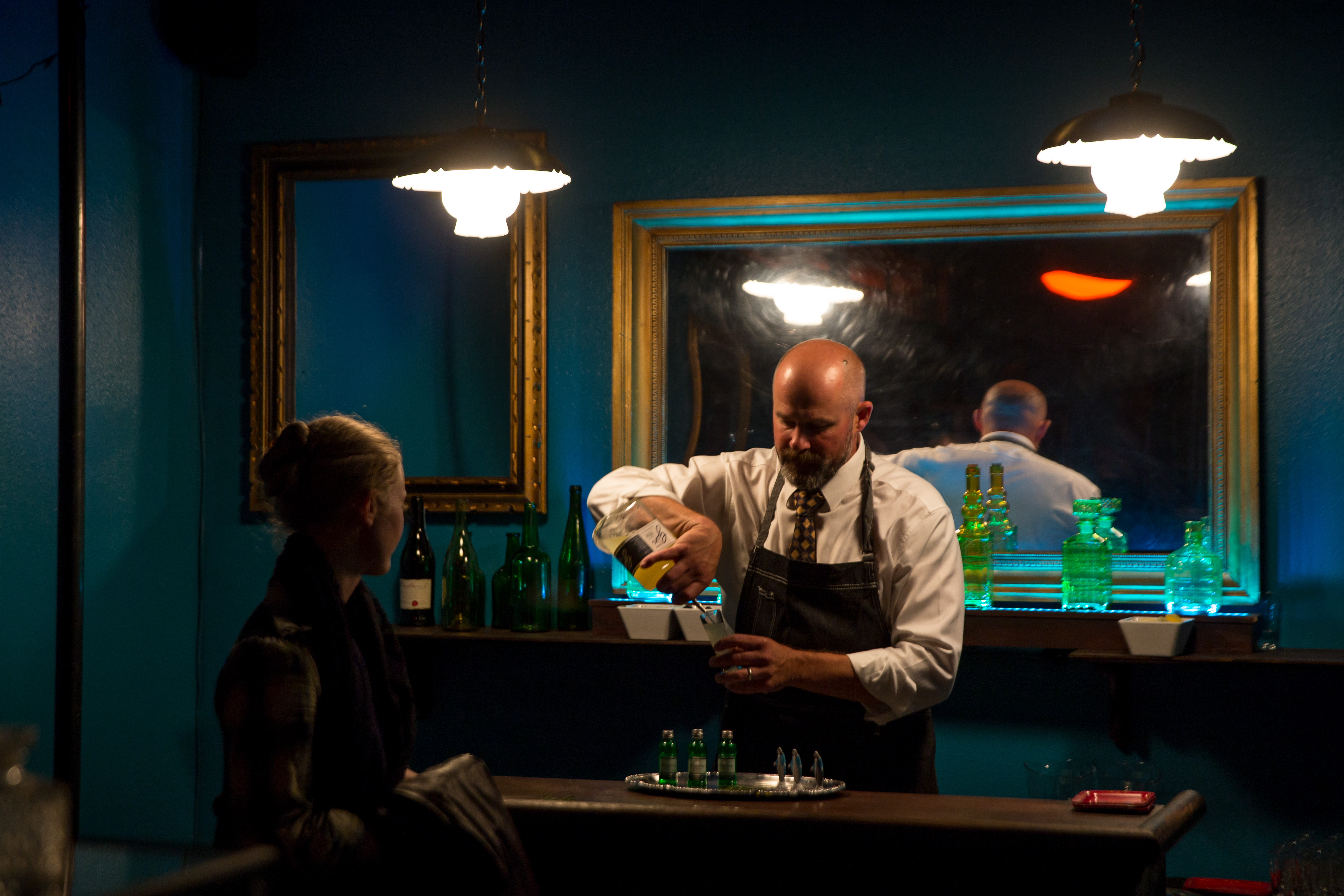 A man pours a drink at a bar while a woman looks on. There is a mirror behind him. ﻿