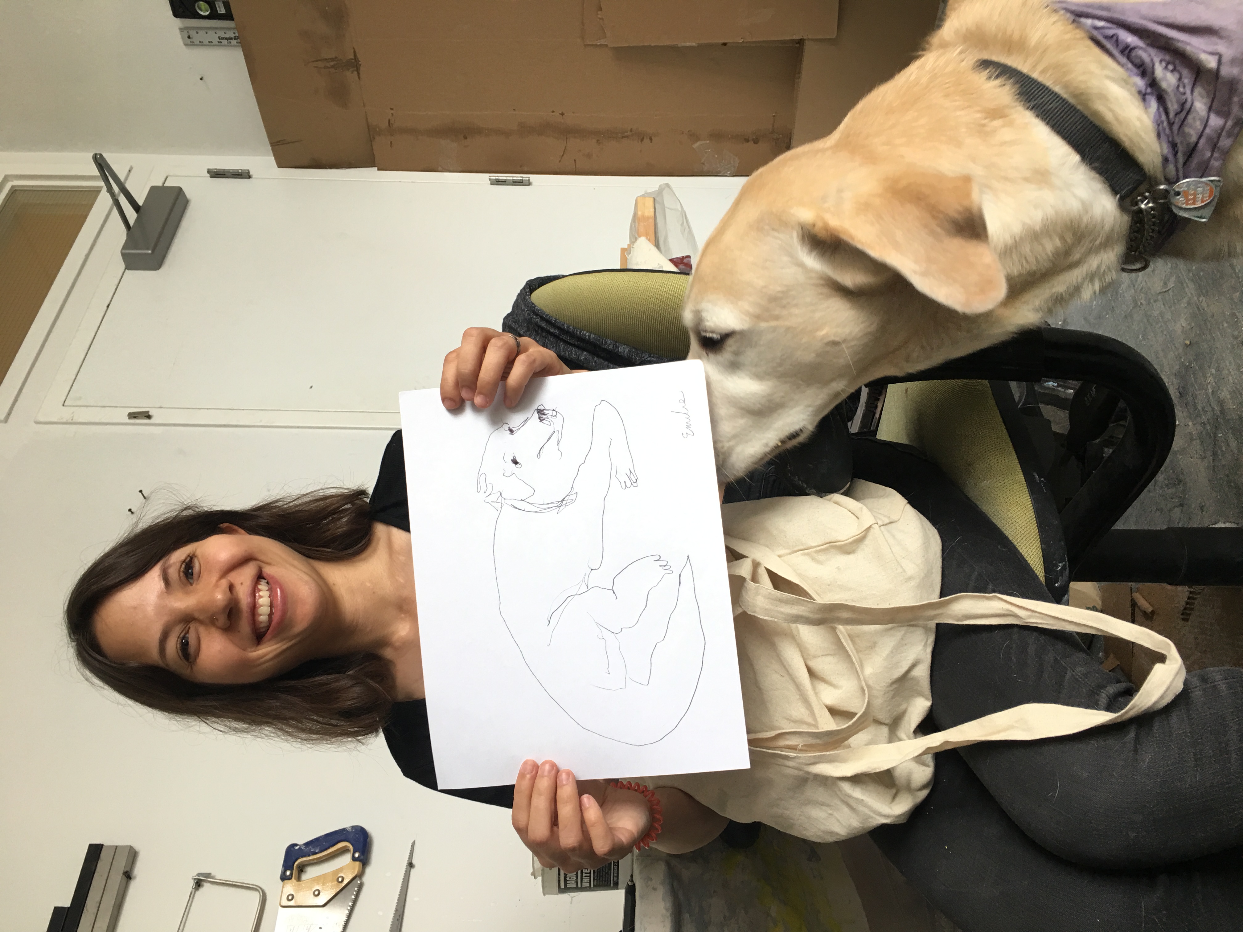 A smiling person with a bob style haircut, holding a drawing of a dog. The dog that appears to be in the drawing is also present in the photo. 