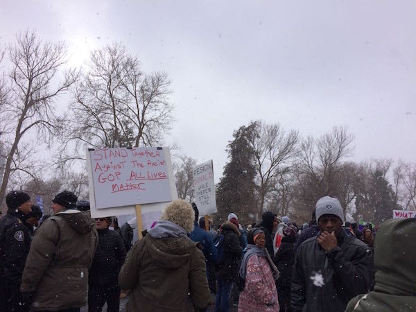 People protest in the snow outdoors. One sign is visible and reads "Stand together against the racist G O P. All lives matter. 