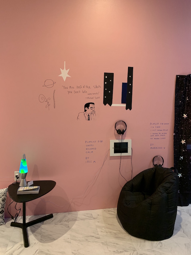 A black beanbag chair lies on the floor next to a lava lamp. A monitor and headphones are mounted to the wall. Text reads “Playlist for happy, relaxed, calm. By Iris M. 