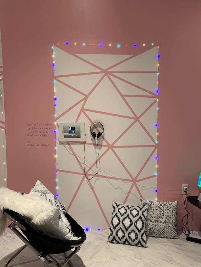 White triangles are painted onto the pink wall. String lights surround the pattern. A monitor and headphones are mounted on the wall. Text reads: acquelin Olivas - This playlist is for when I’m sad and when I’m with friends being in la depre