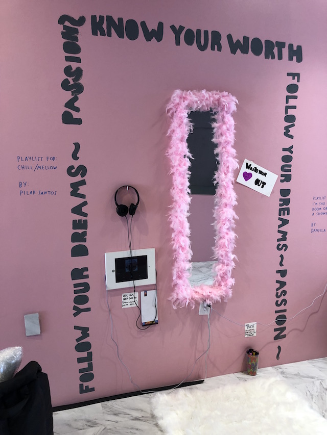A mirror is mounted on the wall next to headphones and a monitor. Pink feather boa surrounds the mirror edges. Black text painted on surrounds the installation reading “Follow your dreams - passion - know your worth- follow your dreams - passion.  Pilar Santos - This playlist is for chill/mellow 